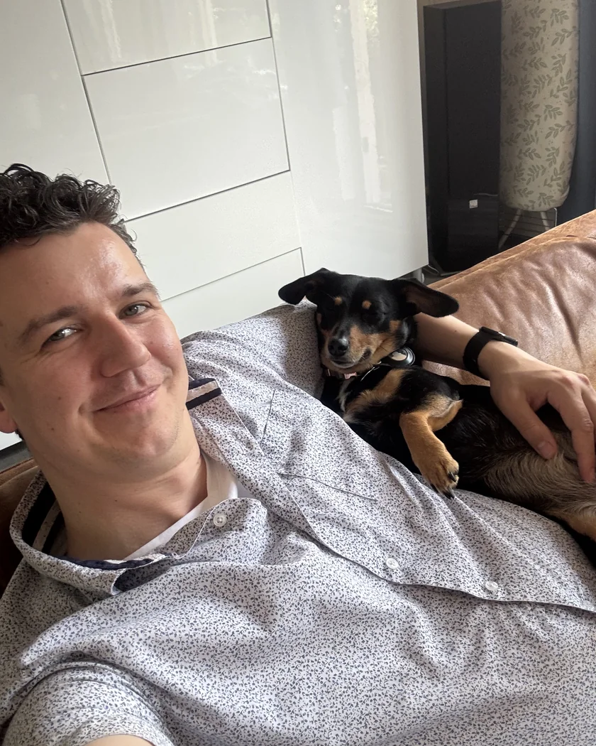 Darius lying back on a brown leather couch and smiling at the camera. Donna, a black and tan dachshund, lies curled up on his chest under his arm with her eyes half closed.