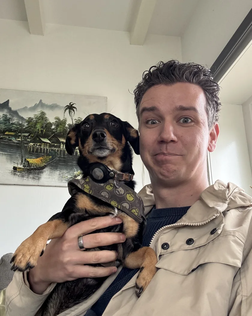 Darius holding Donna, a black and tan dachshund in a grey harness, up to the camera. Both look straight into the lens; he raises his eyebrows, she keeps a straight face.